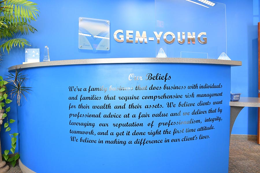 About Our Agency - View of Blue Office Reception Area, Logo Sign, and Gem-Young Insurance & Wealth Advisors Belief System Written on the Front of Desk